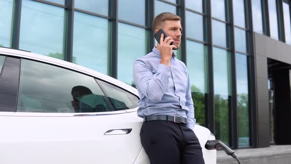 Portrait of Young Handsome European Man Speaking on His Smartphone While Leaning on His Electric Car