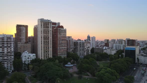 Jib up flying over Barrancas de Belgrano park revealing tall buildings and sunset in background, Bue