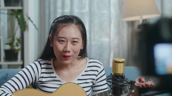 Close Up Of Asian Woman Waving Hand And Speaking To Camera While Live Stream Playing A Guitar