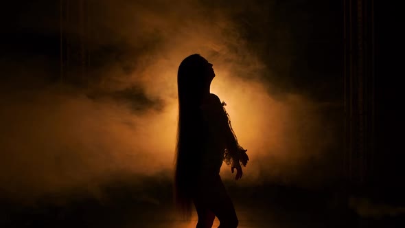 Side View Silhouette of Young Woman Dancing in Dark Smoky Studio Against Background of Yellow Lights