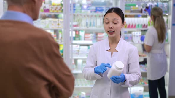 Young Charming Asian Woman Advising Efficient Medication to Senior Caucasian Man Passing Pill Bottle