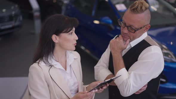 Thoughtful Caucasian Man in Eyeglasses Listening Woman Talking Explaining Vehicle Advantages in Car