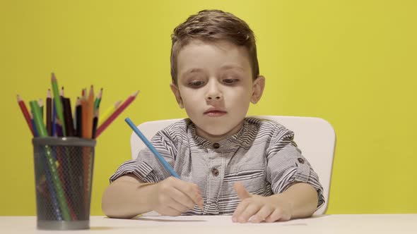 Smiling little boy at the table draw with crayons for mum on yellow background