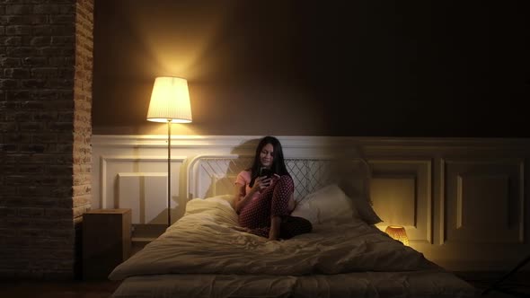 Beautiful girl in pink pajamas sitting in the evening on the bed with the phone