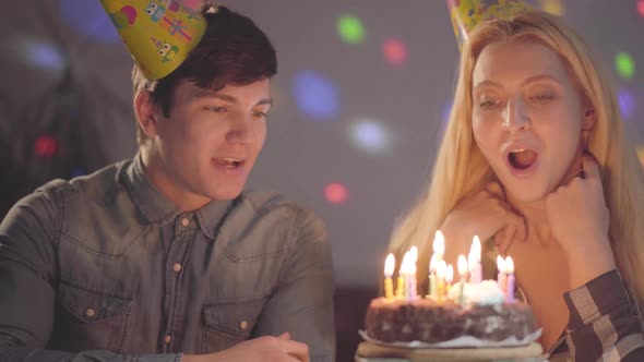 The Blond Girl in Birthday Hat Blowing Out Candles on the Cake, the Young Man Sitting Near