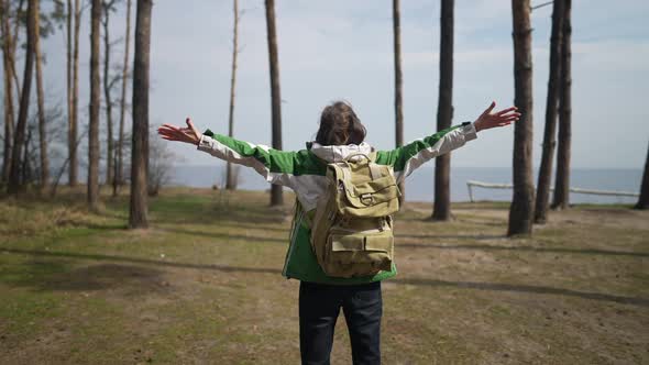 Back View Portrait of Excited Traveler Stretching Hands Standing on Forest Meadow in Front of River