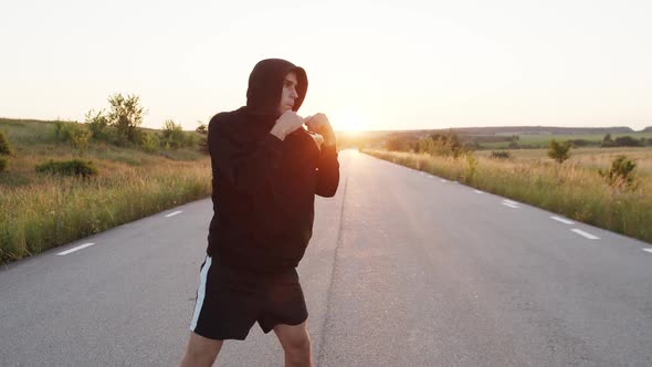 Kickboxer with Hood on Head Training Kicks on Country Road Singly During Sunset