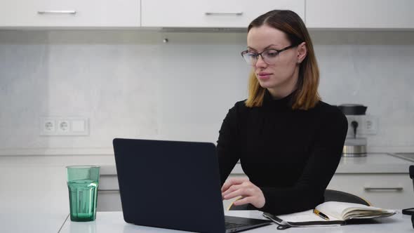 Smart Girl with Glasses Works Behind a Laptop and Makes Notes in a Notebook