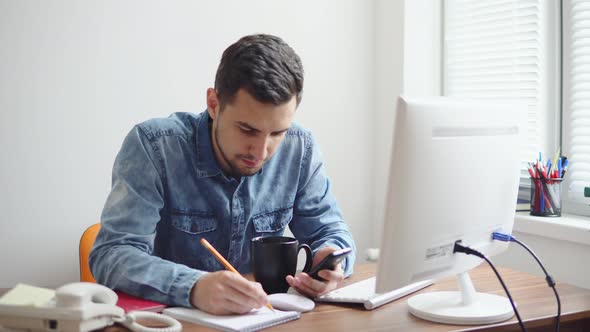 Young Businessman Sitting By the Computer in Stylish Modern Office and Taking Notes Using His Pencil