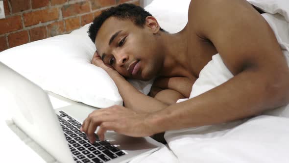 African Man Typing Email on Laptop, Lying in Bed for Rest