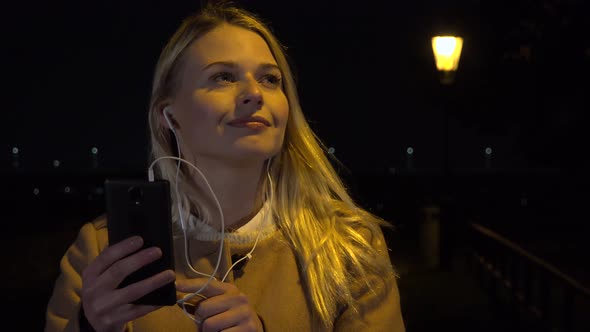 A Young Beautiful Woman Listens To Music on Her Smartphone in an Urban Area at Night