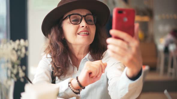 A Mature Stylish Woman in Hat and Glasses Taking a Video Call on a Smartphone