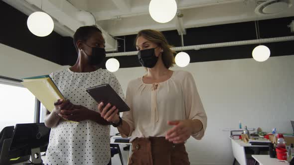 Two diverse female colleagues wearing face masks, walking, and discussing in office