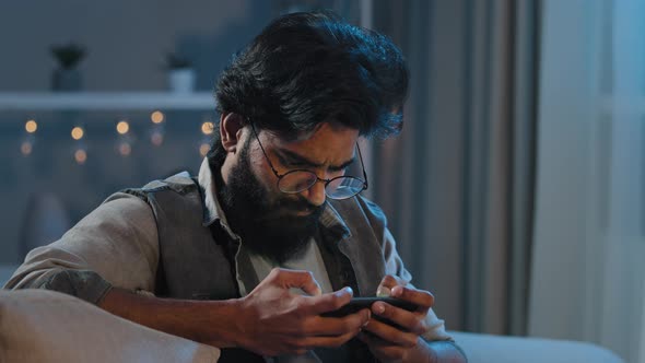Adult Arab Indian Bearded Man with Glasses Sits on Couch at Home in Evening at Night in Dark Late