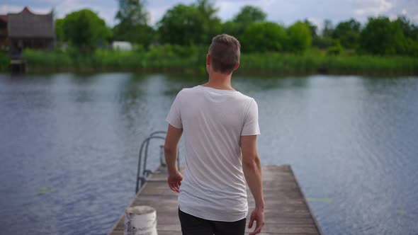 Back View Confident Young Caucasian Man Walking in Slow Motion to River on Wooden Pier Raising Arms