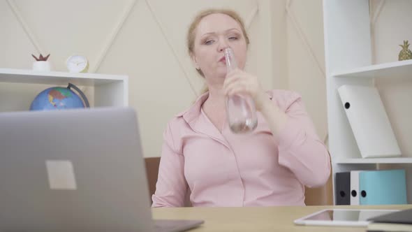 Redhead Senior Woman Drinking Water and Typing on Laptop Keyboard in Office. Portrait of