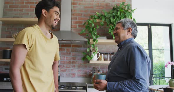 Cheerful biracial father and son talking while toasting coffee mugs in kitchen at home