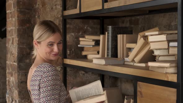 Young attractive woman student turning pages in a book she took from books on the shelves