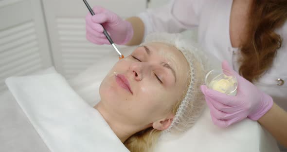 A Young Woman Gets A Procedure From A Beautician.Cosmetologist Making Cosmetic Facial White Mask 