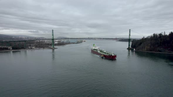 Red barge with pilot boat navigating under Lions Gate bridge with Vancouver city in background, Cana