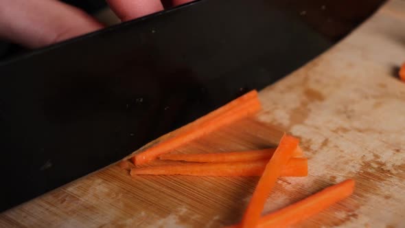 Black knife cuts carrot into thin slices in slow motion, on wooden chopping board.