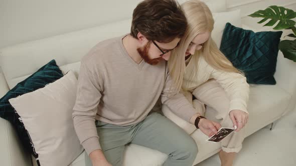 Millennial European Couple Looking at Their Baby Ultrasound Picture While Sitting on a White Sofa in