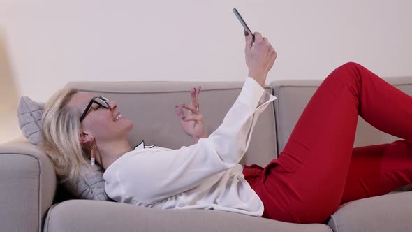 Happy Blonde Woman is Chatting By App in Mobile Phone in Home at Evening Lying on Couch