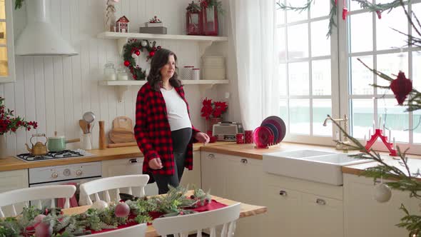 a Young Pregnant Woman in a Red Shirt Stands in a Decorated Kitchen and Looks Out a Large Window