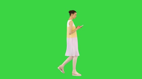 Young Beautiful Girl in Whiteyellow Dress Walks Typing on Mobile Phone on a Green Screen Chroma Key