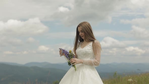 Beautiful Bride in Wedding Dress Stay on the Mountains with Bouquet of Flowers