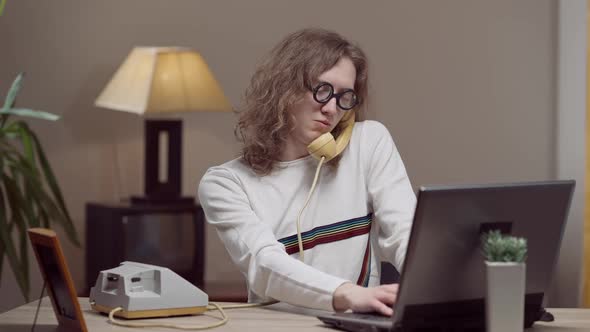 Middle Shot of Concentrated Caucasian Man in Eyeglasses Talking on Retro Telephone and Typing on