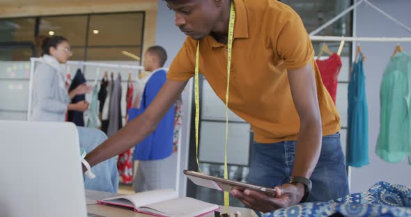 African american male designer standing using tablet and laptop in fashion studio