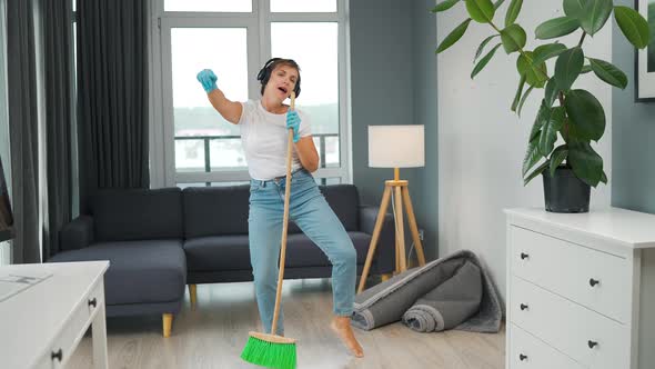 Woman in Headphones Cleans the House and Have Fun Singing with a Broom Like a Star at a Concert