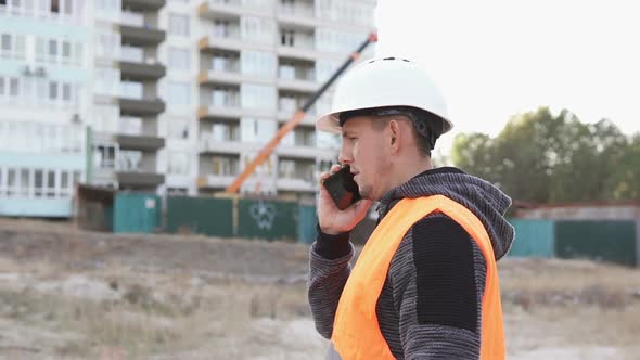 The foreman at the construction site is talking on the phone and checking the drawings.