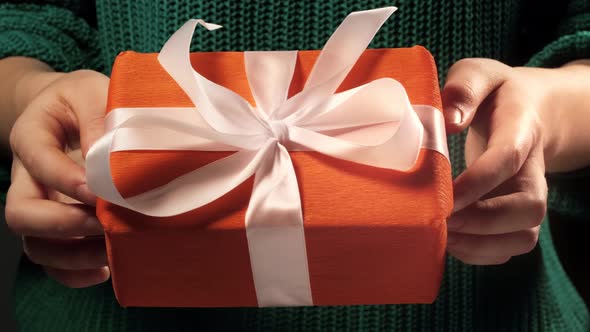 Girl hand holds a gift wrapped in orange paper tied with white ribbon