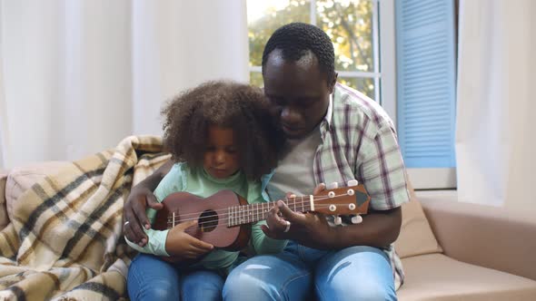 African Toddler Daughter Sitting on Couch Playing Ukulele with Father at Home