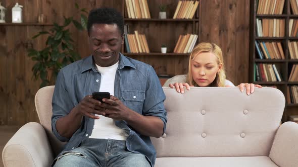 Cheater Man Dating Online with a Smart Phone and Girlfriend is Spying Sitting on a Sofa at Home