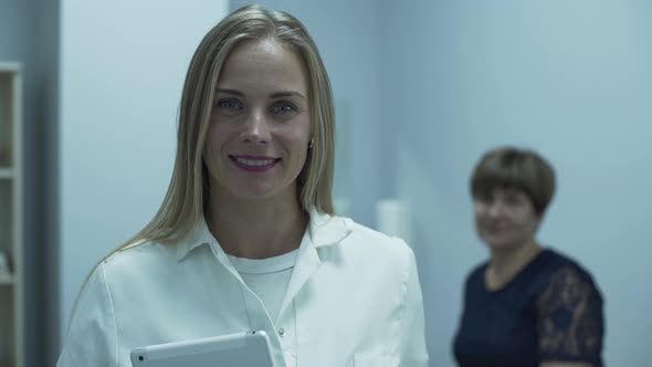 Cute Professional Caucasian Doctor Smiling in Foreground Looking at the Camera While Her Mature