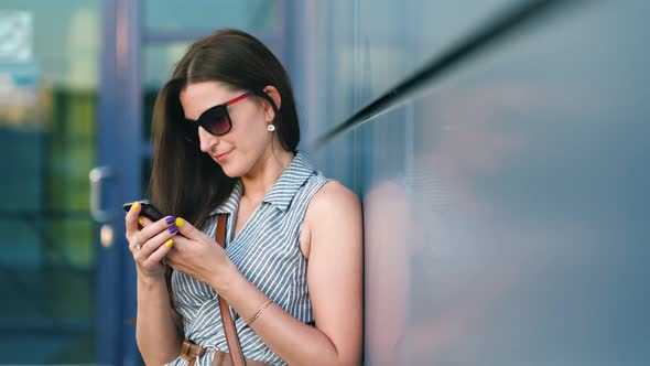 Woman in Sunglasses Using Smartphone Standing Outdoor Leaning Back at Modern Building