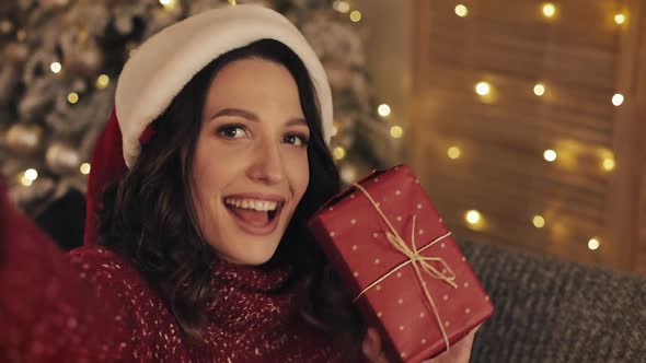 Young Smiling Pretty Brunette Woman Holding Present Box Sitting Near Christmas Tree Shining Lights