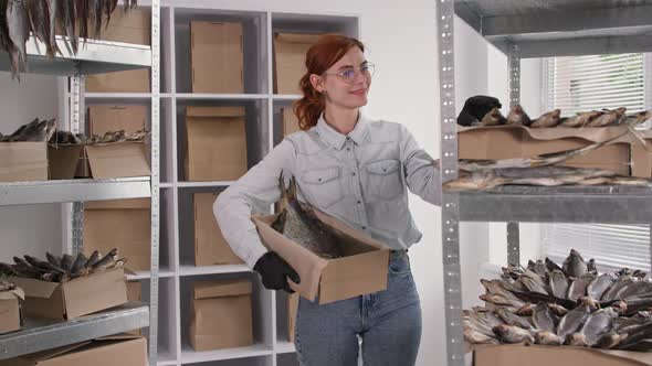 Small Business Smiling Young Woman Wearing Glasses Picks Up Dried Fish From Shelves Into Box for