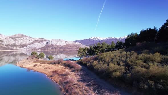 Aerial view through a lake surrounded by nature, trees and a snowy rocky mountain range far in the d