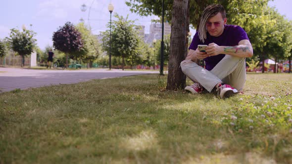 A young man with dreadlocks is resting in the shade of a tree and using a smartphone in the park.