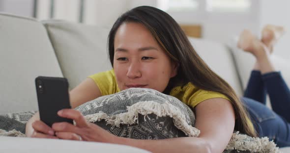 Happy asian woman lying on sofa, resting with smartphone at home