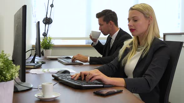 Two office workers, man and woman, sit at desks and work on computers and talk