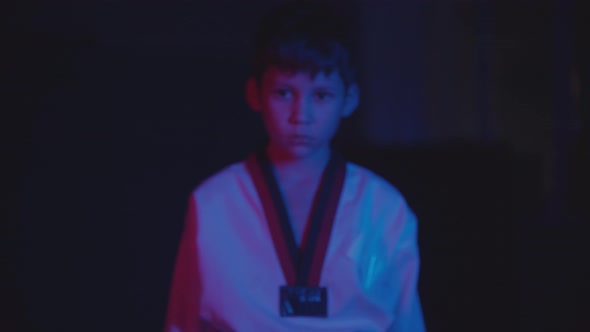 A Little Boy Doing Taekwondo in the Dark with Neon Lighting Walks to the Camera and Showing His