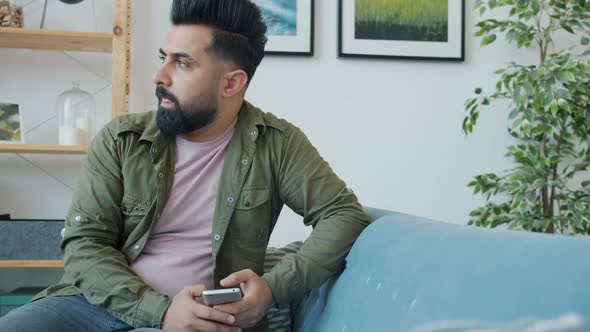 Middle Eastern Man Using Smartphone Getting Bad News Shaking Head Sitting on Couch at Home