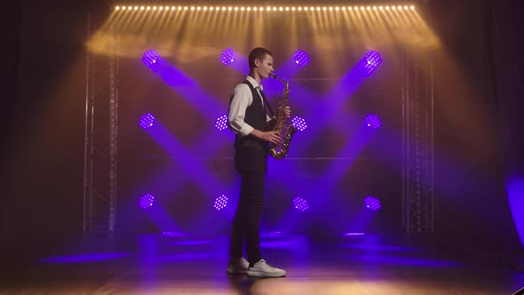 A Young Stylish Guy Plays the Golden Shiny Saxophone in the Blue Spotlights on Stage. Dark Studio