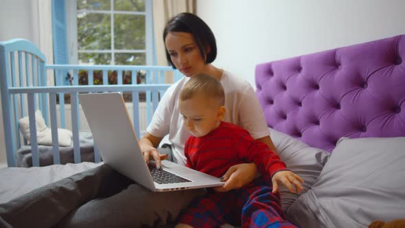 Young Mother and Baby Son Sitting Together at Home on Bed and Using Computer