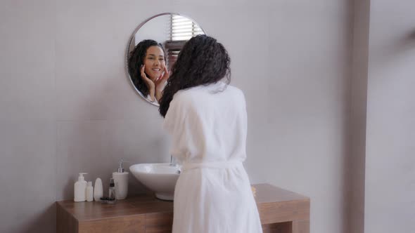 Young Happy Woman Standing in Bathroom Looking in Mirror Touching Face After Applying Cream Enjoys